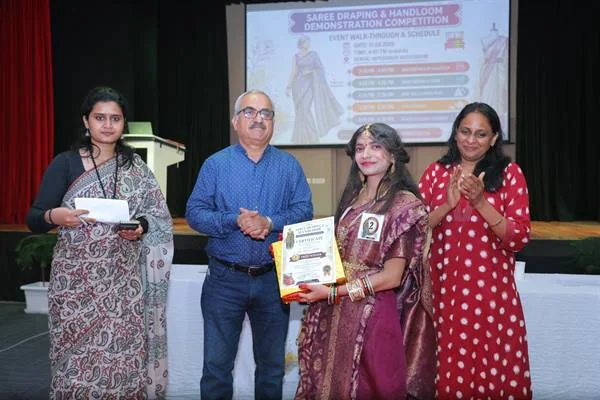 मंच पर चार लोग, एक महिला प्रमाण-पत्र थामे, पीछे प्रतियोगिता की स्लाइड